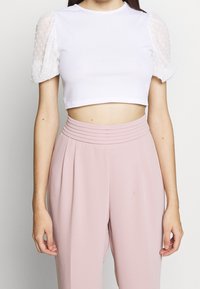 Bílý žebrovaný crop top s průhlednými volánkovými rukávy pokrytými puntíky, spárovaný s světle růžovými kalhotami s vysokým pasem a sklady.