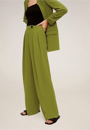 Femme portant un pantalon large vert olive et un blazer assorti sur un top noir, debout avec une main dans la poche et des sandales noires.