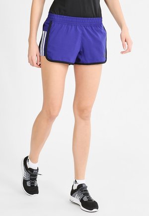 Person iført lilla atletikshorts med sort kant, sorte sneakers og hvide sokker, stående på en hvid baggrund.