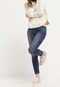 Ljust beige stickad tröja med öppet stickmönster; mörkblå tajta jeans; nude sneakers med vita sulor och snörning; avslappnad outfit.