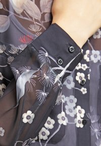 Gros plan d'un chemisier noir transparent à motifs floraux blancs avec des boutons noirs au poignet et sur le devant.