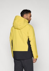 Izoliuota striukė su geltonu viršumi ir juodu apatiniu skyriumi, su kapišonu, lygia tekstūra ir reguliuojamais rankogaliais, kad būtų patogiai priderinama.