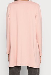 Dlouhý rukáv top v bledě růžové barvě, vyrobený z texturované tkaniny, s kulatým výstřihem a uvolněným střihem, s lemem, který mírně sahá pod boky.