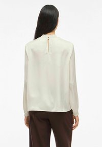 Vrouw met lang, donker haar die een crèmekleurige blouse met lange mouwen en smockmanchetten draagt, met een keyhole achterkant, gecombineerd met donkerbruine broek.