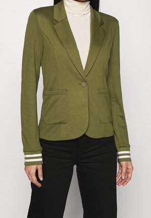 Kobieta ubrana w oliwkowo-zielony żakiet z dwoma przednimi kieszeniami, pojedynczym guzikiem, paskami na mankietach, na białym golfie i czarnych spodniach.