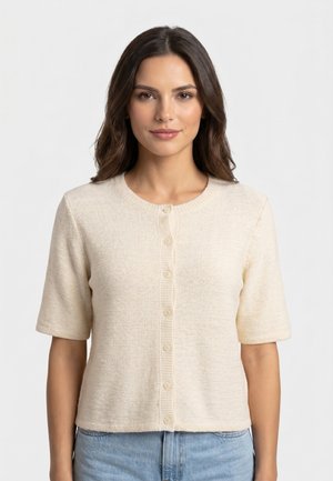 Femme aux longs cheveux bruns portant un pull crème à manches courtes boutonné et un jean bleu clair, debout sur un fond clair uni.
