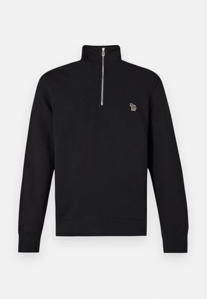 Mladý muž v čiernom svetri na štvrtinový zips, svetlomodrých širokých džínsach a bielych teniskách, stojaci pred jednoduchým bielym pozadím.