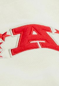 Gesticktes Logo mit einem großen roten "A" auf einem cremefarbenen Hintergrund, mit silbernen Akzenten und einer strukturierten Oberfläche, die auf weichen Stoff hinweist.