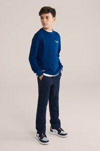 Zils garām piedurknēm krekls ar struktūrētu audumu, balts logo, apvienots ar tumši ziliem sporta biksēm un melniem sporta apaviem ar pelēkiem akcentiem.