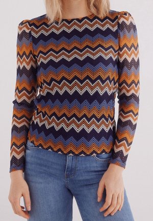 Frau trägt ein langärmeliges Oberteil mit Zickzackmuster in Orange, Marineblau, Blau und Weiß sowie blaue Jeans, stehend vor einem einfarbigen Hintergrund.