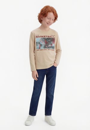 Mladý ryšavý chlapec se usmívá, oblečený v béžovém tričku s dlouhým rukávem a basketbalovým potiskem, modrých džínách a bílých teniskách, stojící s jednou rukou v kapse.
