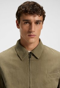 Khaki jakke med en glat tekstur, der har lynlås, stående krave og en enkelt brystlomme.
