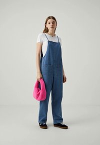 Mlada ženska v modrih denim overallih in beli majici, ki drži svetlo roza torbico, stoji pred enobarvno sivo ozadje.