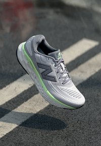 Chaussure de course en maille gris clair avec des accents verts, tige texturée, système de laçage et semelle rembourrée. Présente le logo New Balance.