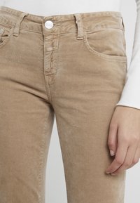 Pantalones de pana beige con un diseño ajustado, que cuentan con un cierre de botón, cinco bolsillos y una textura sutil. Mano visible descansando sobre la tela.