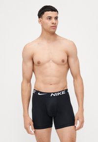 Čierne boxerky Nike vyrobené z hladkej tkaniny majú široký elastický pás s bielymi detailmi loga, ponúkajú pohodlné nosenie a pokrytie do polovice stehna.