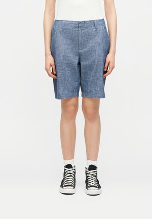 Osoba oblečená v modrých chambray kraťasoch po kolená, bielych ponožkách, čiernych vysokých teniskách a bielom tričku, stojaca s rukami voľne pozdĺž tela.