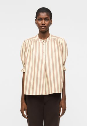 Frau trägt eine beige-weiß vertikal gestreifte Bluse mit kurzen Puffärmeln und dunkle Hose, steht vor einem einfarbigen Hintergrund.