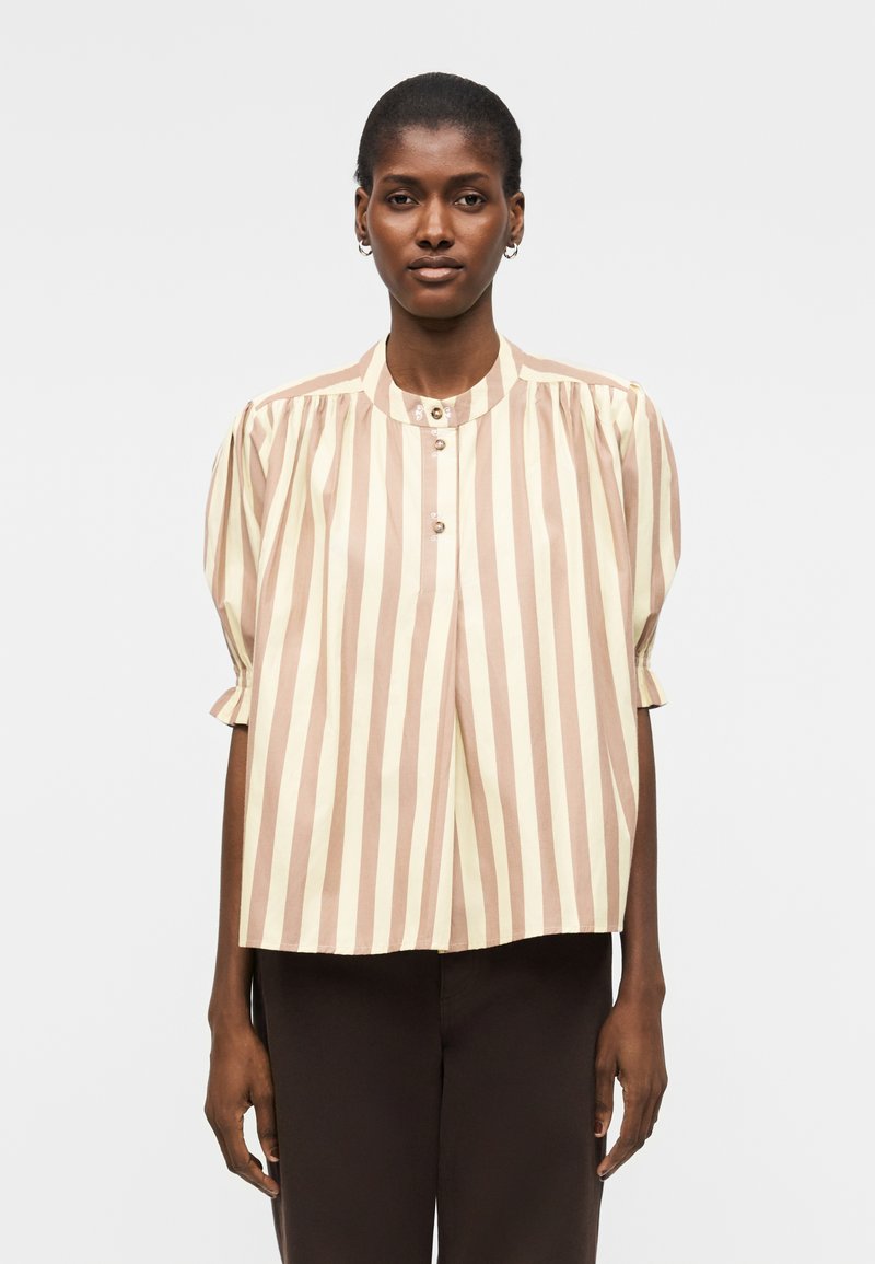 Γυναίκα που φοράει μια μπλούζα με κάθετες μπεζ και λευκές ρίγες με κοντά φουσκωτά μανίκια και σκούρο παντελόνι, στέκεται μπροστά σε απλό φόντο.