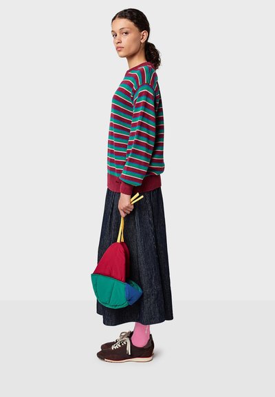 Bunte gestreifte Strickjacke in Rot, Grün und Blau, kombiniert mit einem dunklen Faltenrock. Hält eine kleine gepolsterte, mehrfarbige Tasche. Braune Sneakers.