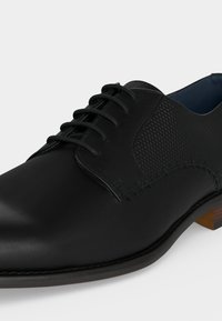 Chaussure habillée en cuir noir avec laçage fermé, panneaux latéraux texturés, détails de couture et un petit talon marron sur fond clair.