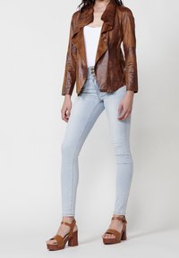Hnědá kožená bunda s límcem, se střihem, který těsně přiléhá k tělu. V kombinaci se světlými modrými skinny džíny a béžovými platformovými sandály.