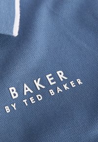 Plava pamučna polo majica s teksturiranom tkaninom; sadrži bijele akcentne boje i otisnuto ime branda "BAKER" u debelom, uzdignutom fontu.