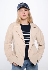 Beige kaksirivinen takki, jossa on läpällinen kaulus, yhdistettynä tummansiniseen raidoitettuun villapaitaan ja tummaan lippalakkiin, jossa on valkoista tekstiä.