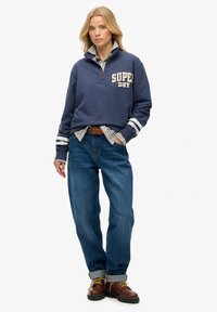 Granatowa bluza z zamkiem błyskawicznym z logo "SUPER DRY", z paskowanymi mankietami, noszona na koszuli w kratę, połączona z luźnymi niebieskimi dżinsami i brązowymi butami.
