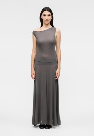 Γυναίκα που στέκεται φορώντας ένα αμάνικο ασύμμετρο γκρι τοπ και μακρύ, ρέον γκρι φούστα με μαύρα παπούτσια πάνω σε απλό φόντο.