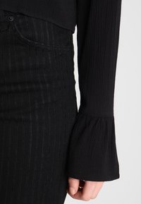 Haut noir à manches longues côtelées avec poignets évasés, associé à un jean noir à rayures fines taille haute, mettant en valeur un tissu texturé.