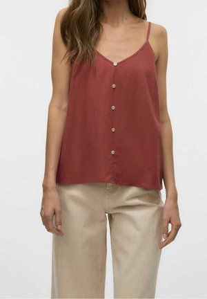 Kobieta ubrana w czerwonobrązową zapinaną na guziki koszulkę na cienkich ramiączkach oraz beżowe szerokie spodnie, na jednolitym tle.