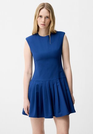 Vrouw met lang blond haar die een mouwloze koningsblauwe jurk draagt met gevoerde schouders en een geplooide rok, staand tegen een witte achtergrond.