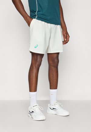 Helle Sportshorts mit Seitentaschen, die ein dezentes Markenlogo und kontrastierende Nähte aufweisen. Kombiniert mit weißen Socken und Schuhen.