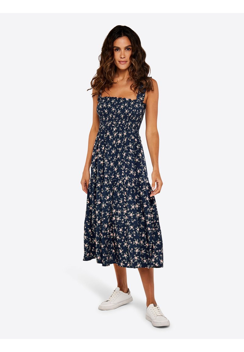 Marineblaues Blumen-Sonnenkleid mit gesmoktem Oberteil, quadratischem Ausschnitt und verstellbaren Trägern. Abgerundet mit einem gestuften Rock und getragen mit weißen Turnschuhen.