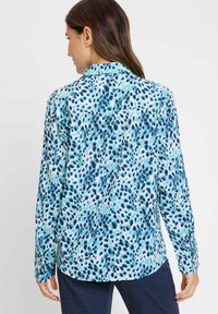 Langarmshirt in lichtblauwe stof met een abstract vlekpatroon in verschillende tinten blauw en marineblauw. Detail van de gebuttonde manchetten zichtbaar.