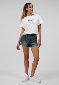 Hvit kortermet t-skjorte med lilla sløyfe-grafikk. Kombinert med denim shorts og hvite sneakers. Enkel, avslappet stil.