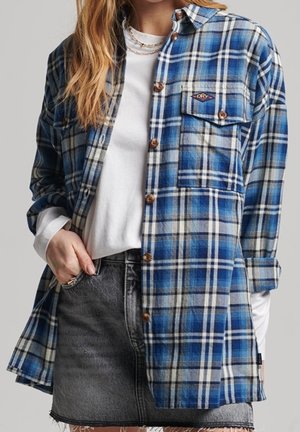 Kobieta ubrana w niebieską i szarą koszulę w kratę na guziki, na białej bluzce z długim rękawem oraz czarną spódnicę dżinsową z frędzlami na brzegu.