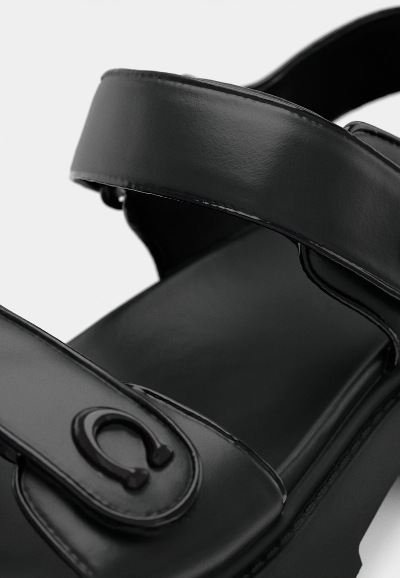 Sandalias negras con dos tiras anchas y brillantes. Presentan un diseño redondeado y un detalle de logo circular prominente en la tira. Textura suave.