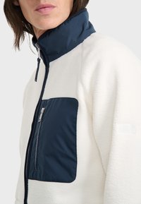 Fleecejacke mit hohem Kragen in Creme, ausgestattet mit einer marineblauen Reißverschlusstasche auf der Brust, kontrastierenden Texturen und einem kleinen Logo-Detail am Ärmel.