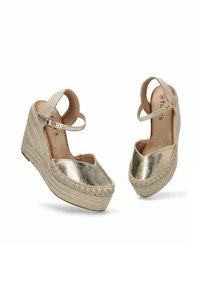 Zapatos de cuña metálicos dorados con suela de yute de espadrille, que presentan una correa en el tobillo entrelazada y una superficie lisa y brillante.