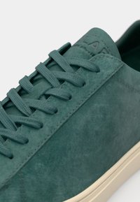 Green suede sneakers with flat laces, featuring a smooth texture, subtle stitching details, and a contrasting cream-colored sole.
