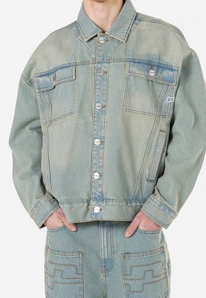 Persoon draagt een lichtblauwe spijkerjasje met knoopsluiting en bijpassende spijkerbroek met grote voorzakken en gestikte ontwerpen.