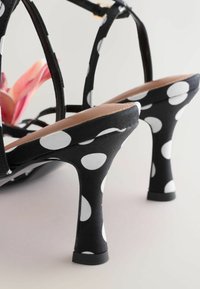 Sandales à talons hauts noires à pois blancs, avec des brides autour de la cheville et un talon arrondi et effilé sur un fond clair.