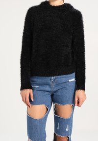 Schwarzer, flauschiger Pullover mit hohem Kragen, kombiniert mit zerrissenen, hellblauen Jeans mit großen Ausschnitten an den Knien.