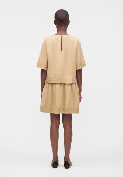 Personne aux cheveux courts portant une robe beige à manches courtes avec des bordures brodées aux manches, à la taille et à l'ourlet, vue de dos, debout sur fond blanc.