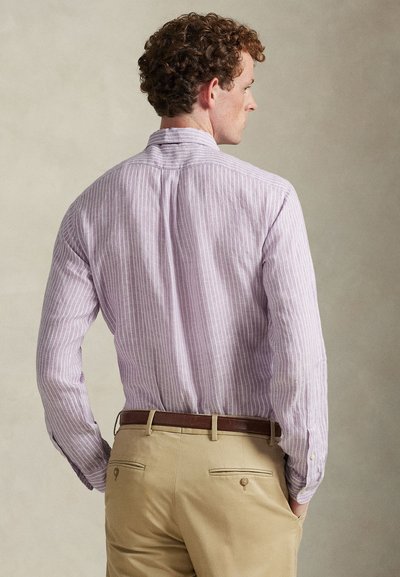 Ανοιχτό μωβ πουκάμισο με ρίγες, μακριά μανίκια, γιακά με κουμπιά και κυρτή βάση, συνδυασμένο με μπεζ παντελόνι και καφέ ζώνη.