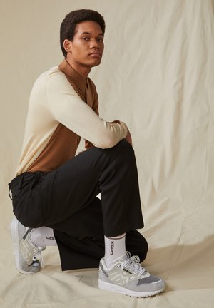 Graue Laufschuhe mit einem Obermaterial aus Wildleder und Mesh, stilvollen Schnürsenkeln und einer dicken weißen Sohle. Das Modell trägt schwarze Hosen und ein zweifarbiges Oberteil.