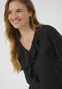 Juoda bluzonė su raukšlėta apykakle, trumpomis rankovėmis ir lygiu, tekstūruotu audiniu. Turi V formos apykaklę ir pritaikytą siluetą.