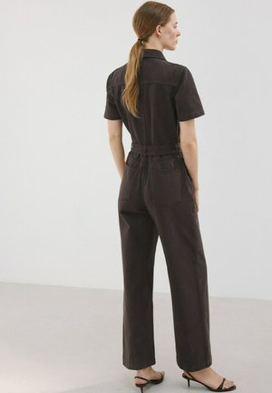 Mujer con el cabello largo recogido en una coleta baja, vistiendo un mono negro de manga corta y sandalias negras de tacón, de pie frente a un fondo claro y liso.